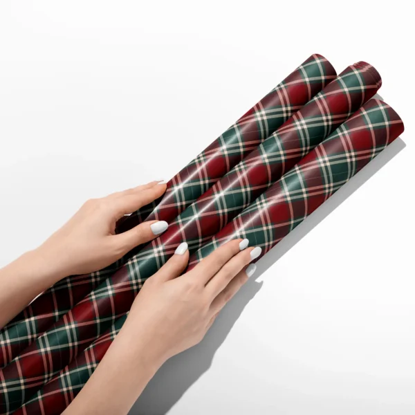 Hands smoothing three rolls of burgundy and green tartan wrapping paper on a white table