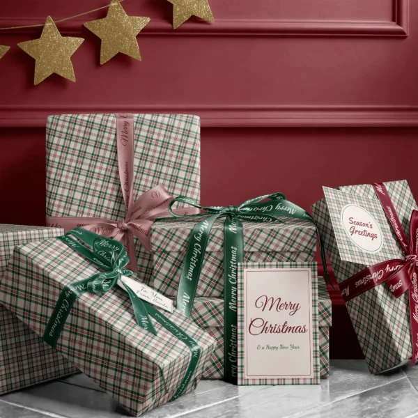 Stack of presents in cream plaid paper with ribbons and a Merry Christmas card by a red wall