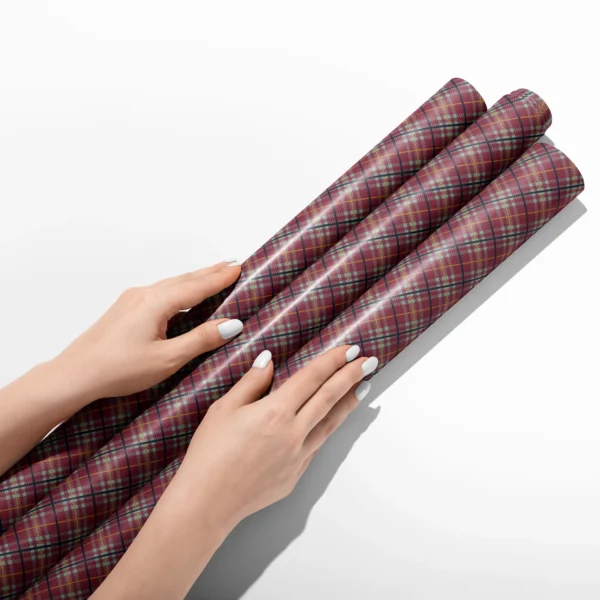 Hands smoothing three rolls of burgundy and sage plaid wrapping paper on a white table