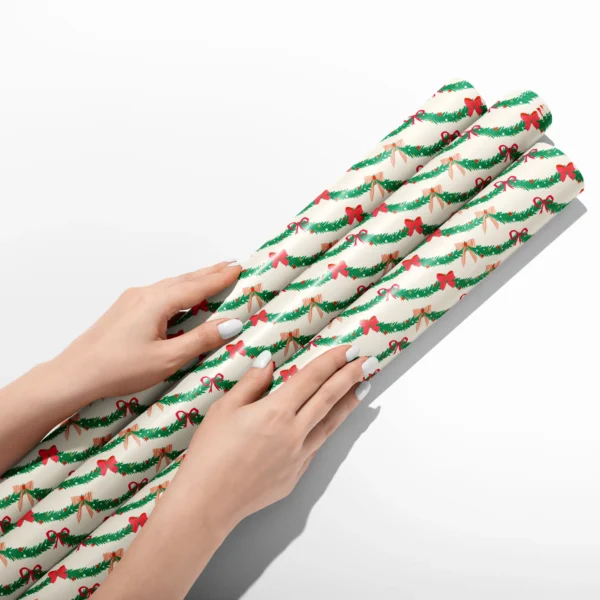 Hands smoothing three rolls of pine garland wrapping paper on a white table