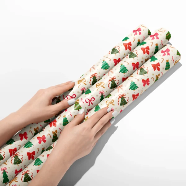 Hands smoothing three rolls of Christmas tree and bow wrapping paper on a white table