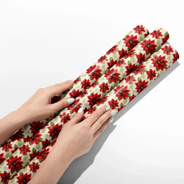 Hands smoothing three rolls of large cream poinsettia wrapping paper on a white table