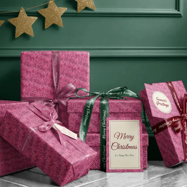 Stack of presents in hot pink spotted paper with ribbons and a Merry Christmas card by a dark green wall