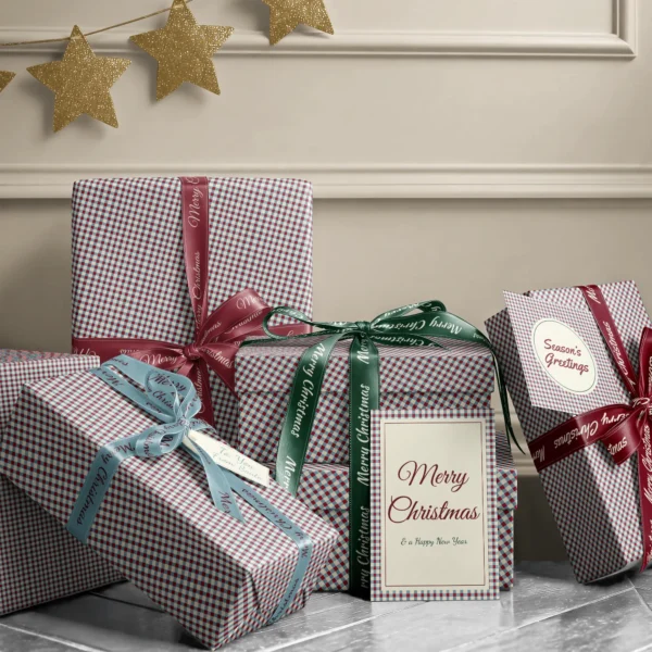 Stack of presents in red and blue gingham paper with ribbons and a Merry Christmas card by a beige wall