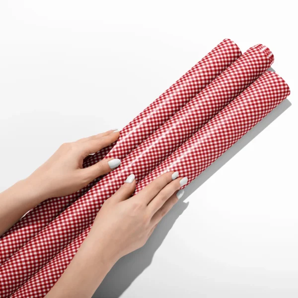 Hands smoothing three rolls of red gingham wrapping paper on a white table