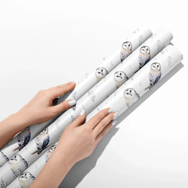 Hands smoothing three rolls of white watercolor owl wrapping paper on a white table